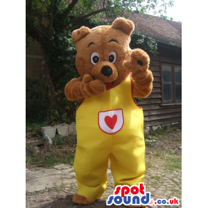 Brown Bear Animal Mascot Wearing Yellow Overalls With A Heart -